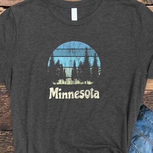 May include: A dark gray t-shirt with a vintage-style graphic of a sunset behind a line of pine trees. The text "Minnesota" is printed below the graphic.