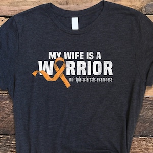May include: A dark gray t-shirt with the text "My Wife Is A Warrior" and an orange ribbon. The text "Multiple Sclerosis Awareness" is below the ribbon.