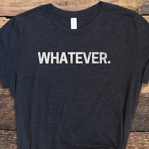 May include: Dark gray t-shirt with the word "WHATEVER." printed in white. The shirt is made of a soft material and has a crew neck. The sleeves are rolled up. The shirt is knotted at the bottom.