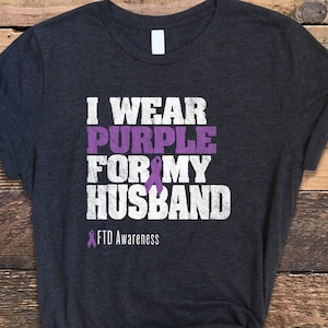 May include: A dark gray t-shirt with the text "I wear purple for my husband" in white with a purple ribbon symbol and the text "FTD Awareness" below.