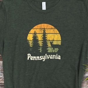 Op de afbeelding: Een donkergroen T-shirt met een vintage-zonnenondergang-afbeelding met een hert en dennenbomen. De tekst "Pennsylvania" staat onder de afbeelding.