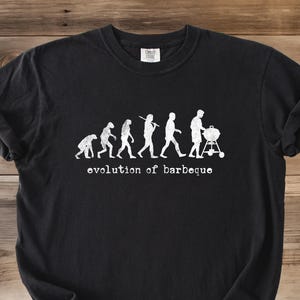May include: Black t-shirt with a white graphic depicting the evolution of humans, culminating in a person pushing a barbecue grill. The text "evolution of barbeque" is below the graphic. The shirt is displayed on a wooden surface.