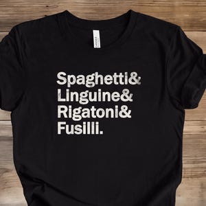 May include: Black t-shirt with white text listing pasta types: Spaghetti, Linguine, Rigatoni, and Fusilli. The shirt is made of a soft material and is displayed on a wooden surface.