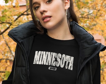 May include: A black long-sleeved shirt with a white distressed graphic that says "Minnesota 1858". The shirt is being worn by a person with a black puffer vest.