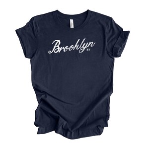 Op de afbeelding: Een marineblauw t-shirt met het woord "Brooklyn" in witte scriptletter. De afkorting "NY" staat eronder. Het t-shirt is gemaakt van zacht materiaal en heeft korte mouwen. Het t-shirt is donkerblauw.