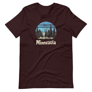 May include: A maroon t-shirt with a vintage-style graphic of a sunset behind a row of pine trees. The graphic is in shades of blue, green, and yellow. The text "Minnesota" is printed below the graphic.