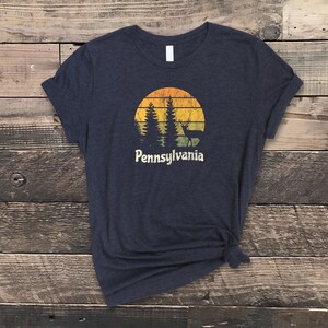 Op de afbeelding: Een donkerblauw t-shirt met een vintage-zonnenondergang-afbeelding met bomen en een hert. De tekst "Pennsylvania" staat onder de afbeelding.