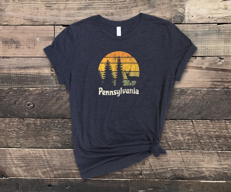 Op de afbeelding: Een donkerblauw t-shirt met een vintage-zonnenondergang-afbeelding met bomen en een hert. De tekst "Pennsylvania" staat onder de afbeelding.