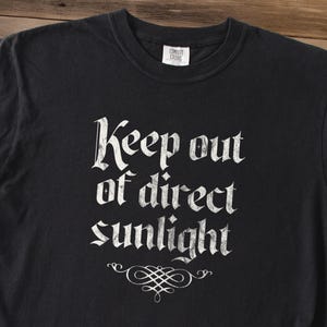 May include: A black short-sleeved t-shirt featuring the white text Keep out of direct sunlight in a distressed gothic font. Below the text is a white decorative flourish. The shirt has a crew neckline and a Comfort Colors brand label at the neck.