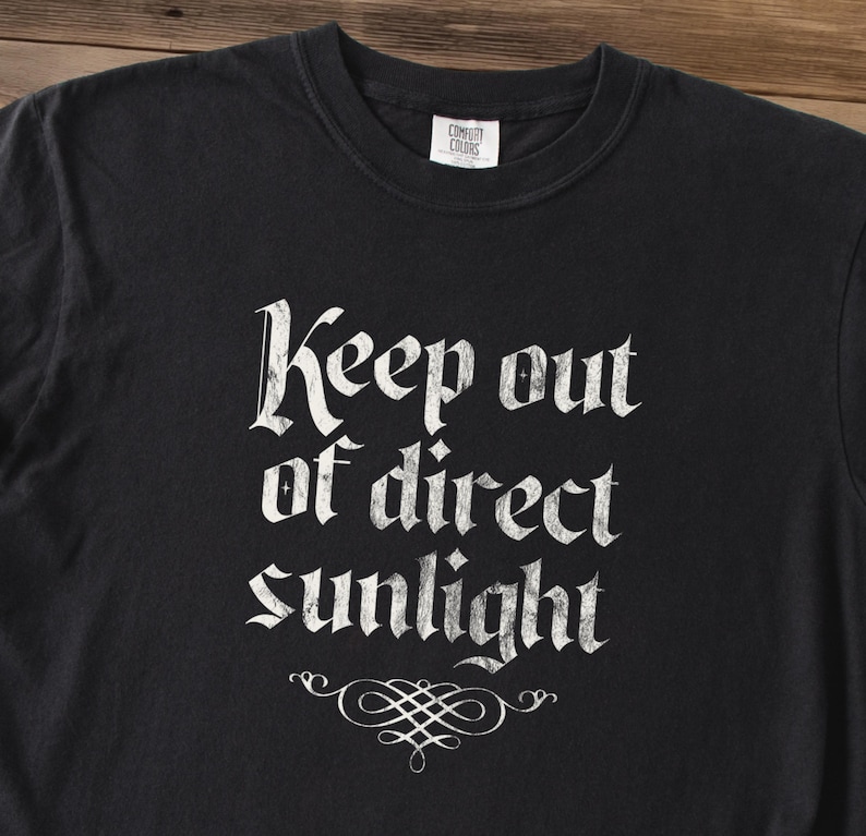 May include: A black short-sleeved t-shirt featuring the white text Keep out of direct sunlight in a distressed gothic font. Below the text is a white decorative flourish. The shirt has a crew neckline and a Comfort Colors brand label at the neck.