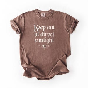 May include: A brown short-sleeve t-shirt with a crew neckline and rolled sleeves. The front features the white text 'Keep out of direct sunlight' in a gothic-style font, positioned above a decorative white flourish graphic centered below the text.