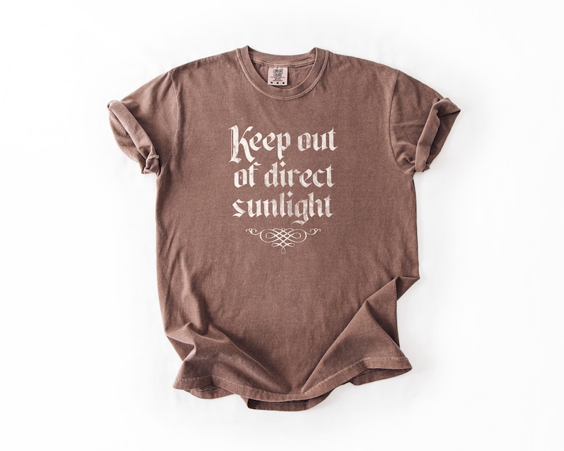 May include: A brown short-sleeve t-shirt with a crew neckline and rolled sleeves. The front features the white text 'Keep out of direct sunlight' in a gothic-style font, positioned above a decorative white flourish graphic centered below the text.