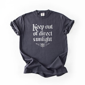 May include: A dark charcoal gray short-sleeve t-shirt featuring the white text Keep out of direct sunlight in a gothic-style font, positioned above a decorative flourish graphic centered on the chest area of the garment.