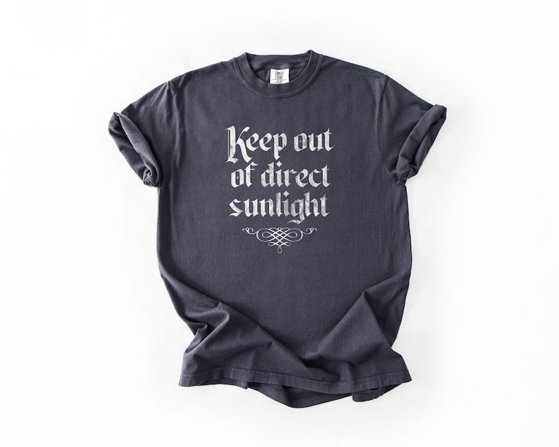 May include: A dark charcoal gray short-sleeve t-shirt featuring the white text Keep out of direct sunlight in a gothic-style font, positioned above a decorative flourish graphic centered on the chest area of the garment.