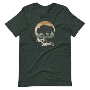 May include: A dark green t-shirt with a vintage-style graphic of a bison silhouette in front of a sunset. The text "North Dakota" is printed below the graphic.