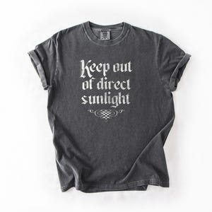 May include: A charcoal gray short-sleeve t-shirt featuring the white text Keep out of direct sunlight in a gothic-style font, centered on the chest above a decorative white flourish. The garment has a crew neckline and rolled sleeves.