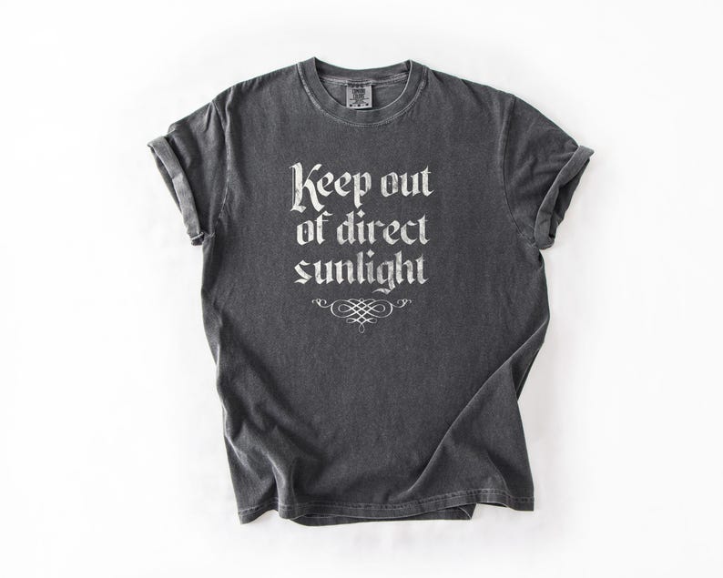 May include: A charcoal gray short-sleeve t-shirt featuring the white text Keep out of direct sunlight in a gothic-style font, centered on the chest above a decorative white flourish. The garment has a crew neckline and rolled sleeves.