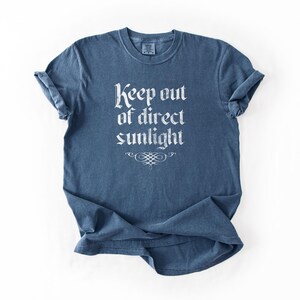 May include: A blue short-sleeve t-shirt with a crew neckline. The front features white text that reads Keep out of direct sunlight in a gothic-style font, positioned above a decorative white flourish graphic centered on the chest area.