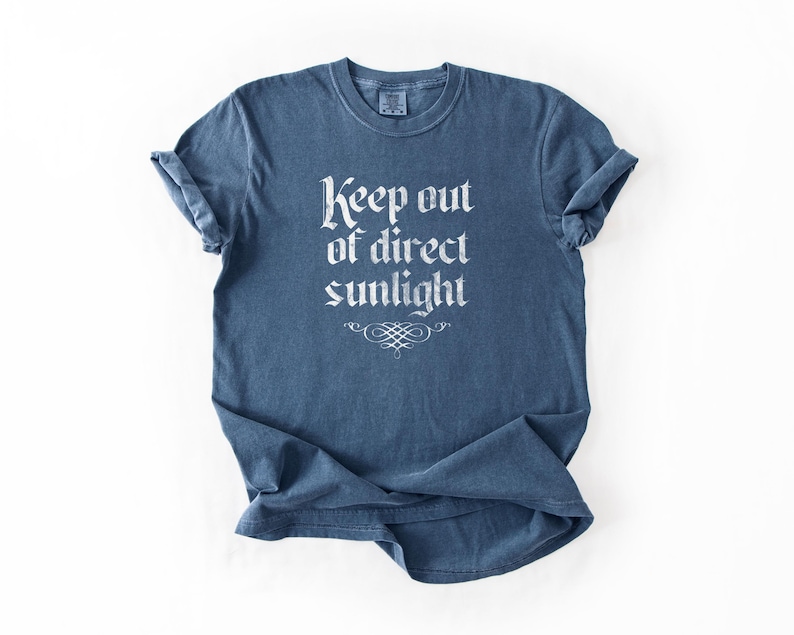 May include: A blue short-sleeve t-shirt with a crew neckline. The front features white text that reads Keep out of direct sunlight in a gothic-style font, positioned above a decorative white flourish graphic centered on the chest area.