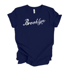 Op de afbeelding: Een marineblauw t-shirt met het woord "Brooklyn" in witte letters op de voorkant. De tekst "NY" staat onder het woord "Brooklyn". Het shirt is gemaakt van een zacht materiaal.