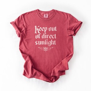May include: A short-sleeved, crew-neck t-shirt in a faded red color. The center of the chest features the white text "Keep out of direct sunlight" in a gothic-style font, positioned above a small, decorative, symmetrical scrollwork flourish.