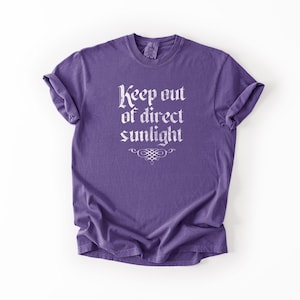 May include: A purple short-sleeve t-shirt featuring the white text "Keep out of direct sunlight" printed in a gothic-style font on the chest, accompanied by a small decorative flourish centered directly below the text.