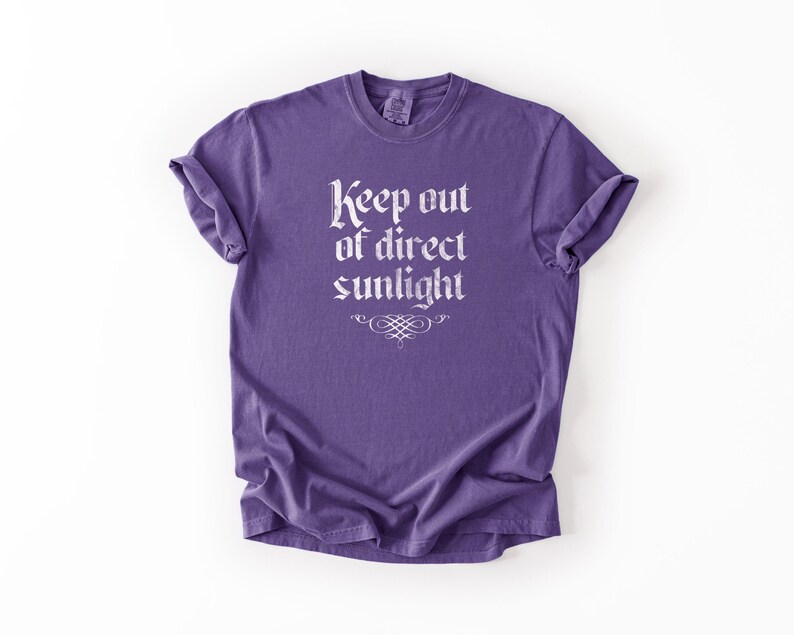 May include: A purple short-sleeve t-shirt featuring the white text "Keep out of direct sunlight" printed in a gothic-style font on the chest, accompanied by a small decorative flourish centered directly below the text.