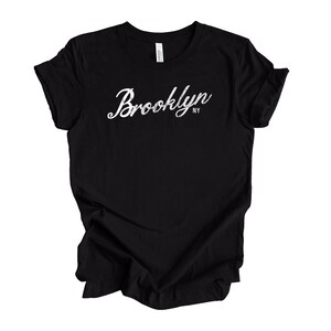 Op de afbeelding: Zwart T-shirt met het woord "Brooklyn" in witte letters op de voorkant. De tekst bevat "NY" in kleinere letters onder het hoofdword. Het shirt is gemaakt van een zacht materiaal.