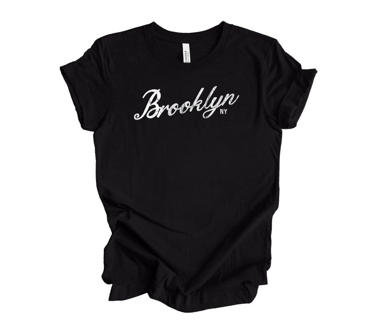 Op de afbeelding: Zwart T-shirt met het woord "Brooklyn" in witte letters op de voorkant. De tekst bevat "NY" in kleinere letters onder het hoofdword. Het shirt is gemaakt van een zacht materiaal.