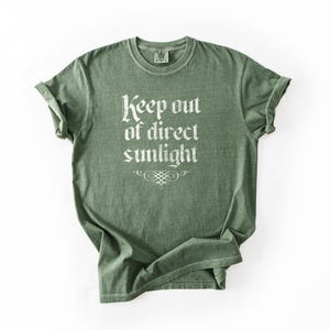 May include: A sage green short-sleeve crew neck t-shirt featuring the white text "Keep out of direct sunlight" printed in a gothic-style font on the chest, accompanied by a small decorative flourish graphic centered below the text.