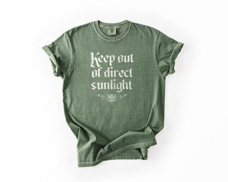 May include: A sage green short-sleeve crew neck t-shirt featuring the white text "Keep out of direct sunlight" printed in a gothic-style font on the chest, accompanied by a small decorative flourish graphic centered below the text.