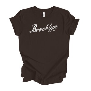 Op de afbeelding: Een donkerbruin t-shirt met het woord "Brooklyn" in witte sierlijke letters over de borst. De letters "NY" staan eronder. Het shirt is gemaakt van een zacht materiaal.
