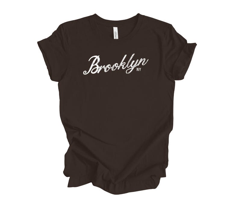Op de afbeelding: Een donkerbruin t-shirt met het woord "Brooklyn" in witte sierlijke letters over de borst. De letters "NY" staan eronder. Het shirt is gemaakt van een zacht materiaal.
