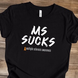 May include: Black t-shirt with the text "MS SUCKS" in white, with an orange awareness ribbon and the words "multiple sclerosis awareness" in white. The shirt is made of a soft material.