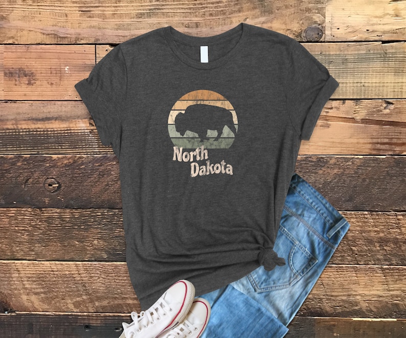May include: A dark heather grey t-shirt with a vintage-style graphic of a bison silhouette in front of a sunset. The text "North Dakota" is printed below the graphic.