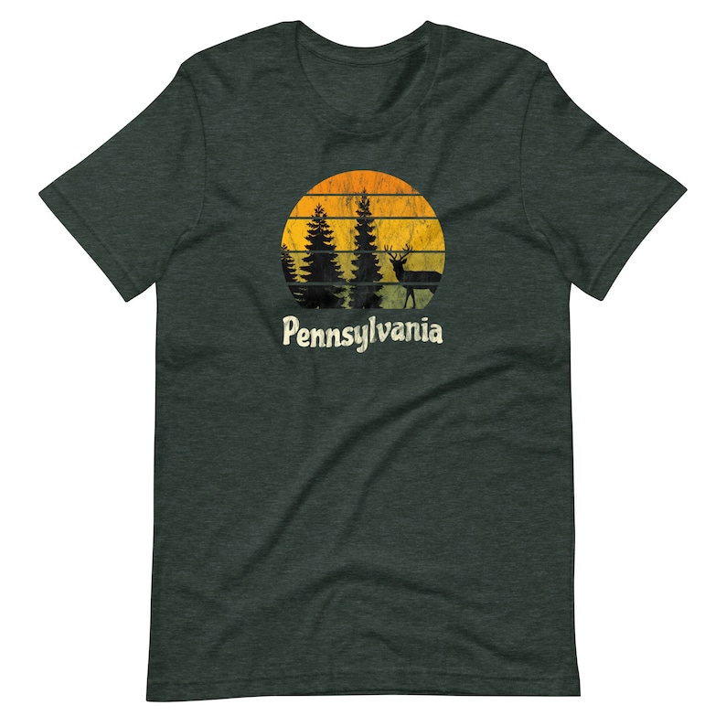 Op de afbeelding: Een donkergroen T-shirt met een vintage-stijl afbeelding van een zonsondergang met bomen en een hert. De tekst "Pennsylvania" is onder de afbeelding gedrukt.