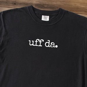 May include: Black t-shirt with the phrase "uff da" in white, centered on the front. The shirt has a crew neck and a tag that says "Comfort Colors" at the neckline.