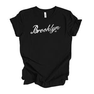 Op de afbeelding: Zwart t-shirt met het woord "Brooklyn" in witte scriptletters. De letters "NY" staan onder het woord "Brooklyn". Het t-shirt is gemaakt van een zacht materiaal.