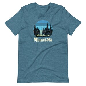 May include: A blue, heather grey t-shirt with a vintage-style graphic of a sunset behind a row of pine trees. The graphic is in shades of blue, yellow, and green. The text "Minnesota" is printed in a retro font below the graphic.