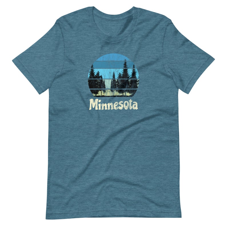 May include: A blue, heather grey t-shirt with a vintage-style graphic of a sunset behind a row of pine trees. The graphic is in shades of blue, yellow, and green. The text "Minnesota" is printed in a retro font below the graphic.