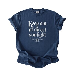 May include: A navy blue short-sleeve t-shirt featuring the white text Keep out of direct sunlight printed in a gothic-style font on the chest, accompanied by a decorative flourish graphic centered directly below the text.