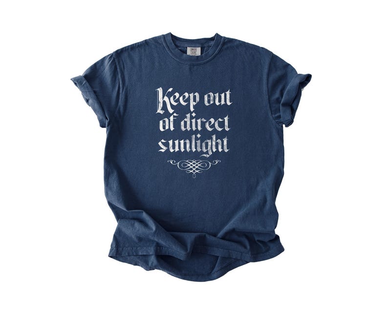 May include: A navy blue short-sleeve t-shirt featuring the white text Keep out of direct sunlight printed in a gothic-style font on the chest, accompanied by a decorative flourish graphic centered directly below the text.