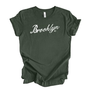 Op de afbeelding: Donkergroen t-shirt met het woord "Brooklyn" in witte sierlijke letters. Daaronder staat de afkorting "NY". Het t-shirt is gemaakt van een zacht materiaal en heeft korte mouwen. Het is een casual kledingstuk.