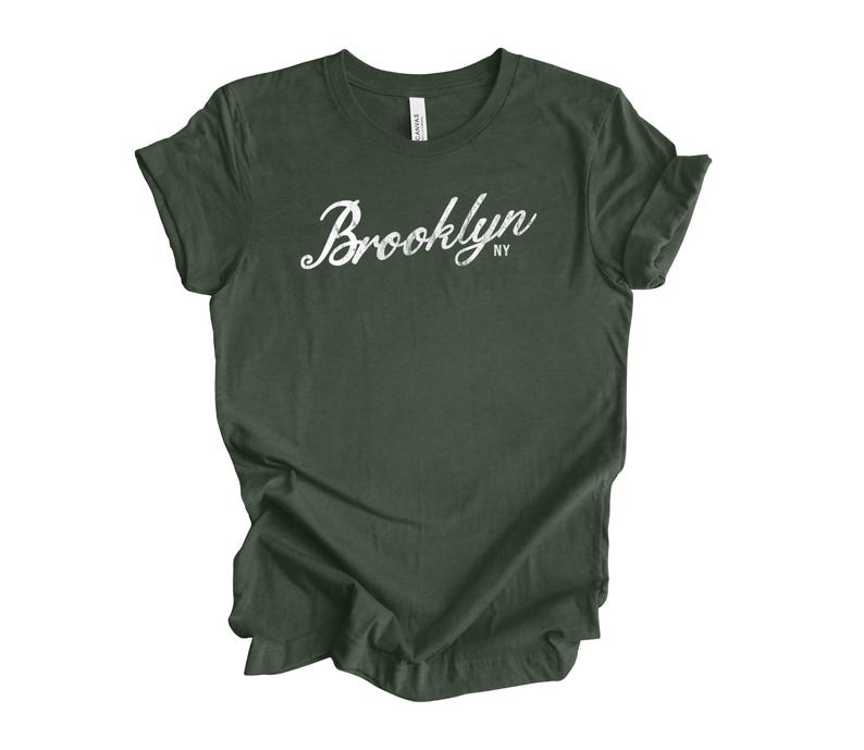 Op de afbeelding: Donkergroen t-shirt met het woord "Brooklyn" in witte sierlijke letters. Daaronder staat de afkorting "NY". Het t-shirt is gemaakt van een zacht materiaal en heeft korte mouwen. Het is een casual kledingstuk.