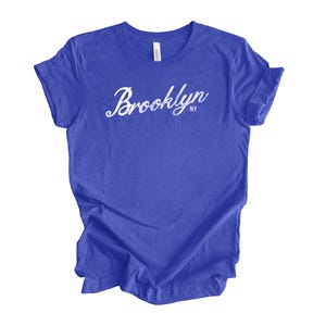 Op de afbeelding: Een koningsblauw t-shirt met het woord "Brooklyn" in witte cursieve letters. De tekst "NY" staat onder het woord "Brooklyn". Het t-shirt is gemaakt van een zacht materiaal en heeft korte mouwen.