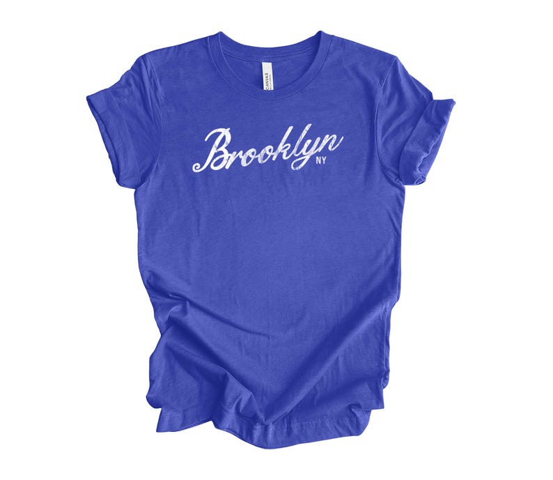 Op de afbeelding: Een koningsblauw t-shirt met het woord "Brooklyn" in witte cursieve letters. De tekst "NY" staat onder het woord "Brooklyn". Het t-shirt is gemaakt van een zacht materiaal en heeft korte mouwen.
