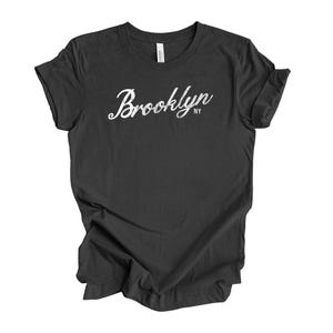 Op de afbeelding: Een donkergrijs t-shirt met het woord "Brooklyn" in een wit scriptlettertype. De letters "NY" staan onder het woord "Brooklyn". Het t-shirt is gemaakt van een zacht materiaal en heeft korte mouwen.