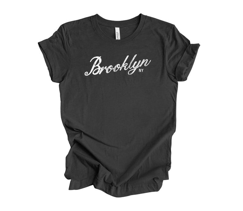 Op de afbeelding: Een donkergrijs t-shirt met het woord "Brooklyn" in een wit scriptlettertype. De letters "NY" staan onder het woord "Brooklyn". Het t-shirt is gemaakt van een zacht materiaal en heeft korte mouwen.