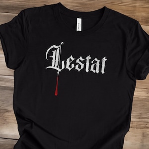May include: Black t-shirt with the name "Lestat" in white gothic lettering. A single drop of red, resembling blood, hangs from the bottom of the letter "t". The shirt is made of a soft material.