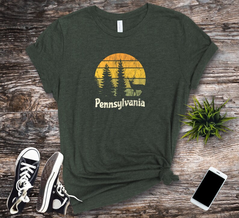 Op de afbeelding: Een donkergroen T-shirt met een vintage-zonnenondergang-afbeelding met een hert en dennenbomen. De tekst "Pennsylvania" staat onder de afbeelding.